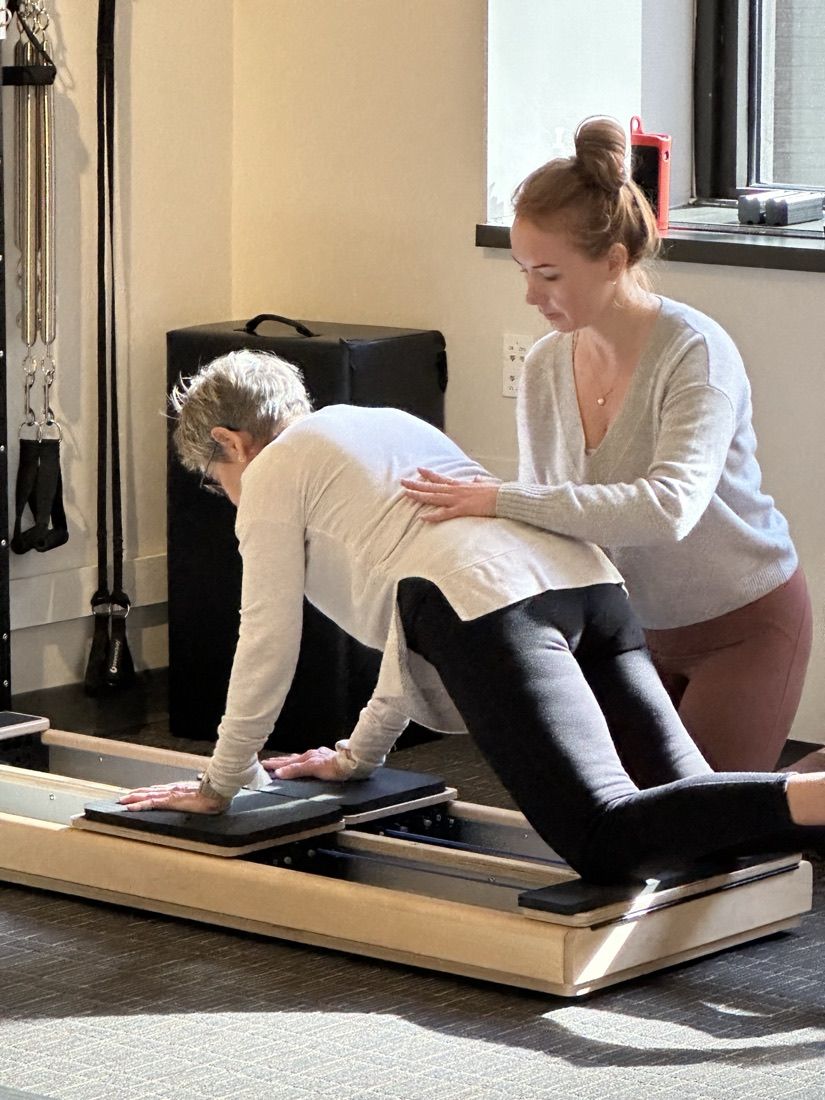 A coach guiding an older client through a balance and strength sequence on the reformer